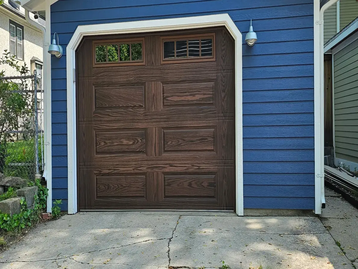 Brown carriage style garage door with window inserts on a blue sided home